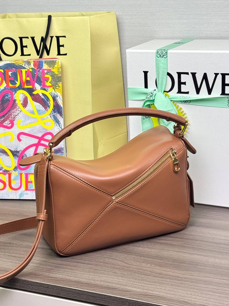 🎈【周年限定】 小号纳帕羊皮𝗙𝗲𝗮𝘁𝗵𝗲𝗿𝗹𝗶𝗴𝗵𝘁 𝗣𝘂𝘇𝘇𝗹𝗲手袋👜 ——山核桃色到货❤️ 尺寸：𝟮𝟰-𝟭𝟬.𝟱-𝟭𝟲.𝟱𝗰𝗺 编码：061610【配全套礼盒】 ✅延续经典老款𝗣𝘂𝘇𝘇𝗹𝗲做法，全新𝗣𝘂𝘇𝘇𝗹𝗲内外采用柔软轻盈的纳帕羊皮材质，为手袋注入随性慵懒的风格。 ✅该款饰有旋转卡扣，铰链式拉链头，一个外部拉链后袋，一个内部插袋，以及可拆卸式的𝗟𝗢𝗘𝗪𝗘烫金或烫银标签，饰有隐秘口袋和气球骰子，底部长方形金属脚钉。搭配可拆卸可调节长肩带，可肩背、斜挎或手提。